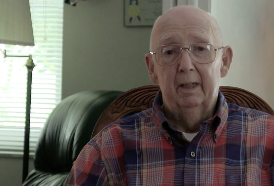 Elder man with plaid shirt speaking to a camera.